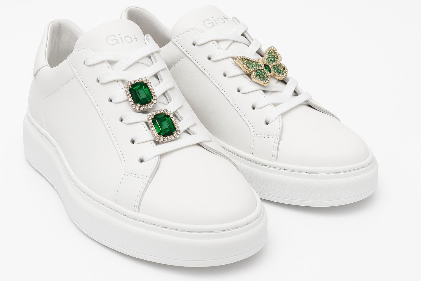 Sneakers Bianca Gio+ con accessori infilati nei lacci a dx una farfalla con pietre oro e verdi a sx 2 pietre quadrate verdi rifinite con zirconi chiari che girano intorno alla pietra rettangolare
